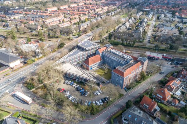 Luchtfoto van een groot kantoorgebouw aan de Lage Naarderweg in Hilversum, omgeven door woonwijken en geparkeerde auto's.